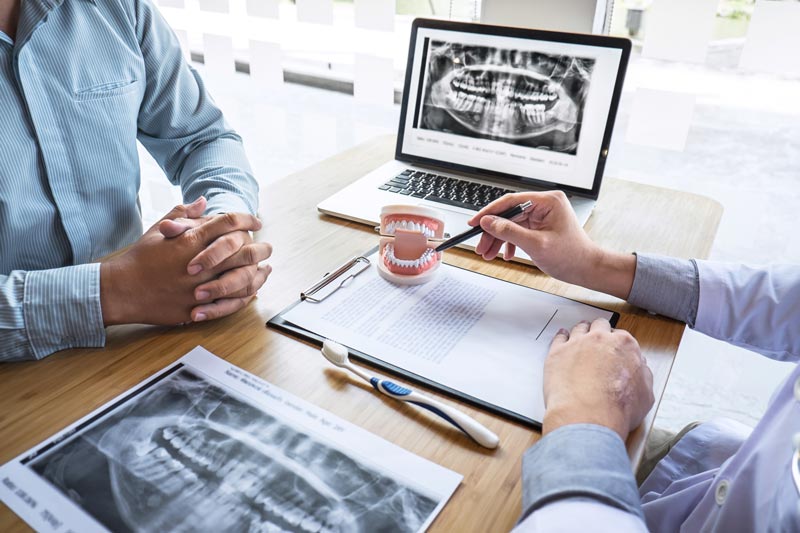 The image shows a dental professional using a digital X-ray machine with a patient sitting across from them, discussing a dental treatment plan.