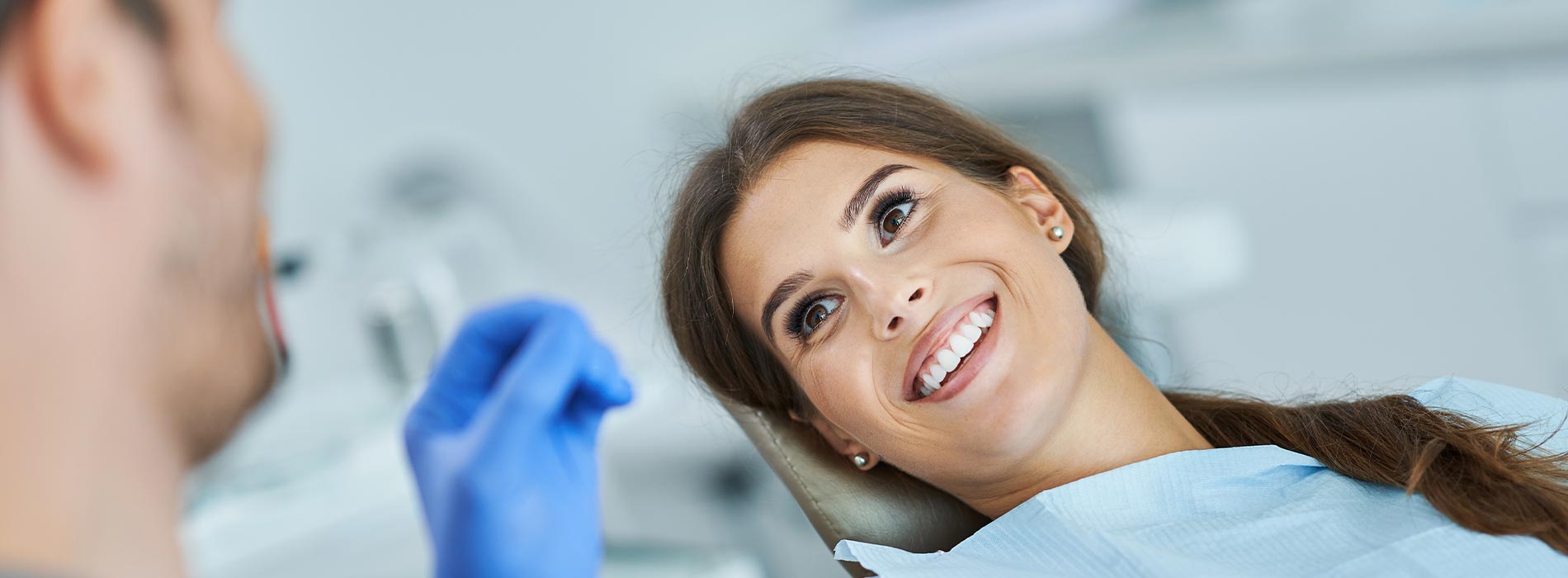 The image shows a woman smiling at the camera while seated in a dental chair, with a professional-looking person standing behind her, both wearing medical masks and gloves, indicating a dental care setting.