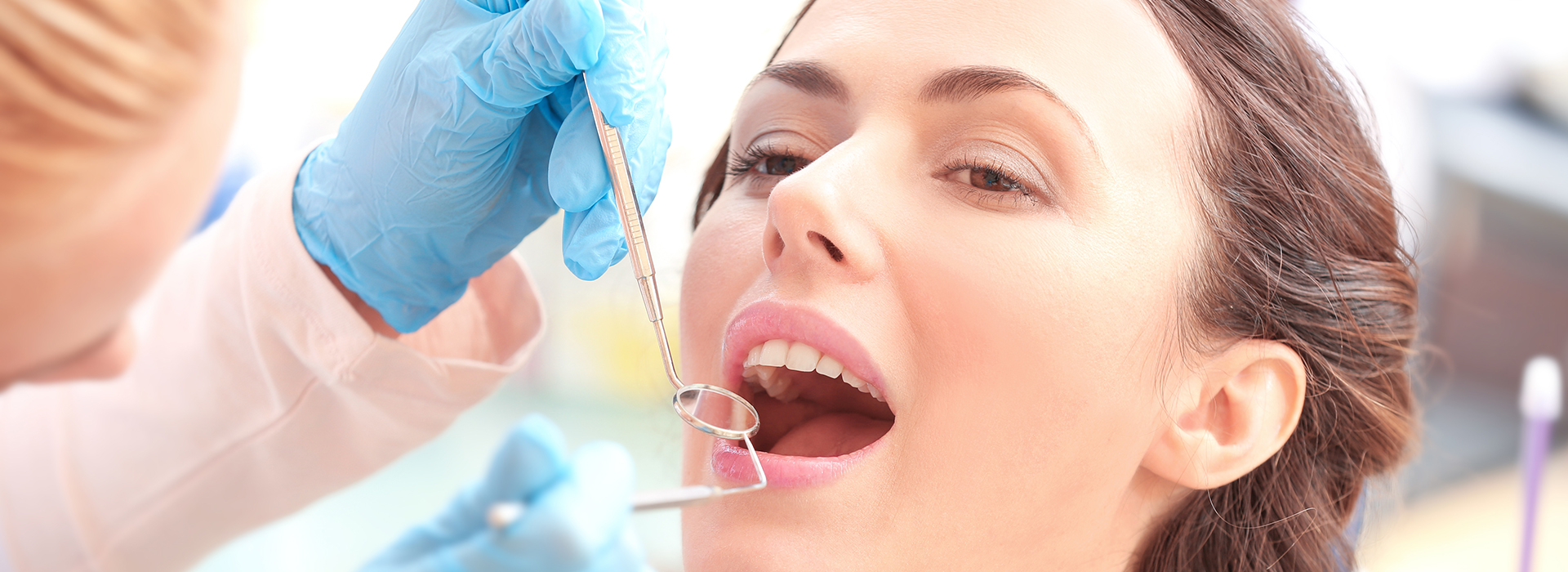 This is a photograph featuring a woman seated with her mouth open wide, receiving dental care from someone out of frame who appears to be a dentist, with a close-up view of the dental instruments being used on her teeth.