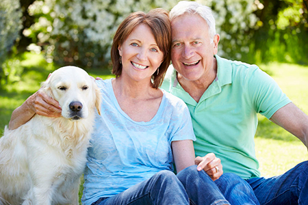 A man and woman sitting on grass with a dog between them, smiling at the camera.