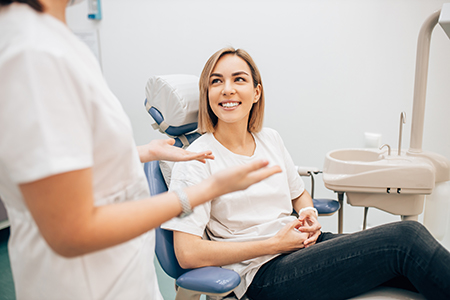 A woman sitting on a dental chair with a smile, being attended by a dentist who is smiling and gesturing towards her.